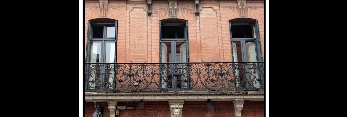 Façade toulousaine en briques roses aux fenêtres cintrées et volets, balcon filant en fer forgé sur consoles sculptées