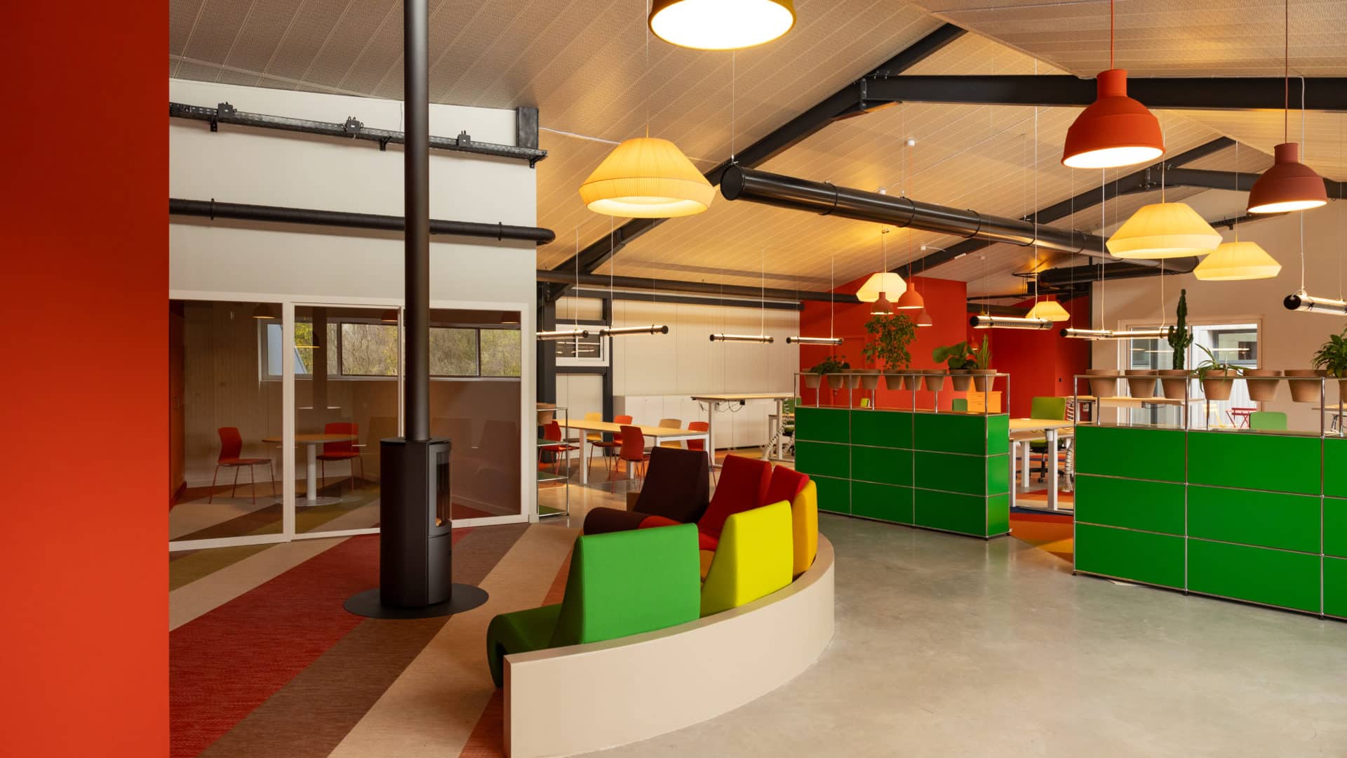 Bureaux Huck Occitania : plateau ouvert coloré avec banquette courbe multicolore, rangements vert vif, poêle à bois et charpente métallique noire apparente sous un plafond bois