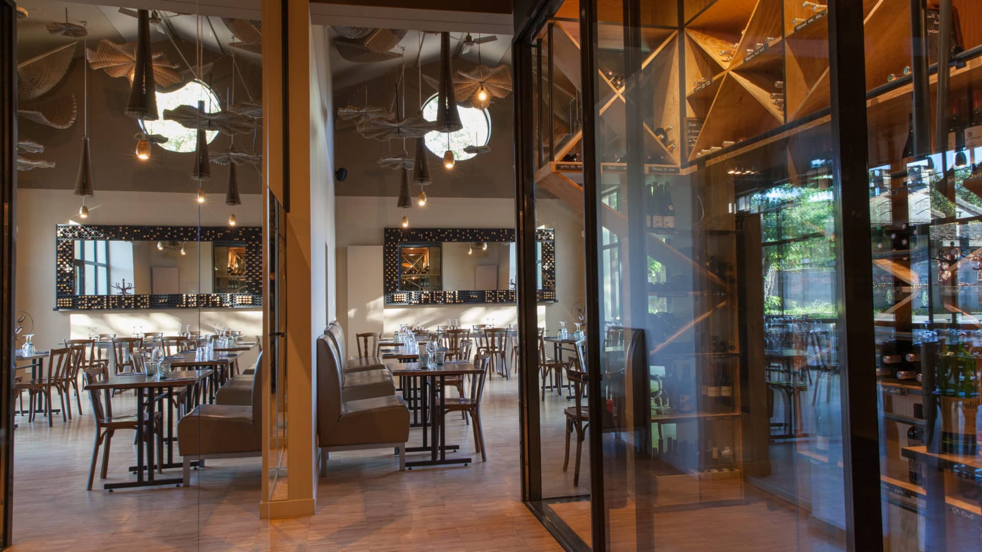 Salle du restaurant Label Braise : tables en bois et banquettes, suspensions tressées et fenêtre ronde, et cave à vin vitrée au casier bois en diagonale