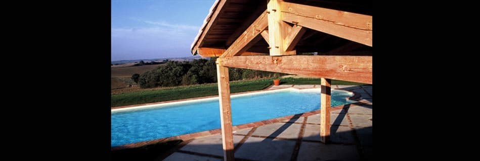 Terrasse couverte en bois surplombant la piscine et les collines cultivées du Gers au coucher du soleil