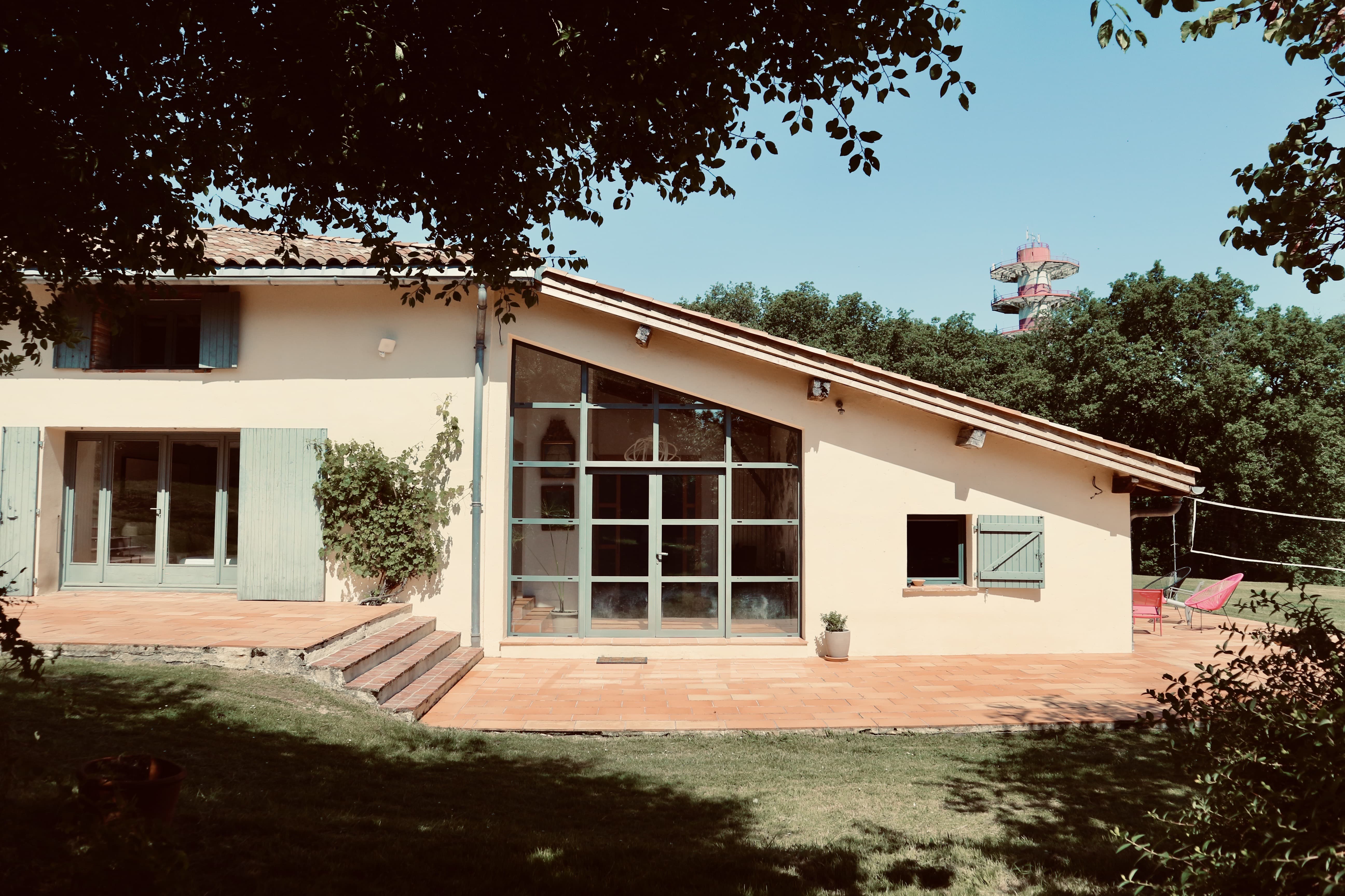 Extérieur de la maison : extension en appentis à grande verrière encadrée de volets vert pâle, terrasse en tomettes et jardin arboré sous la lumière d'été