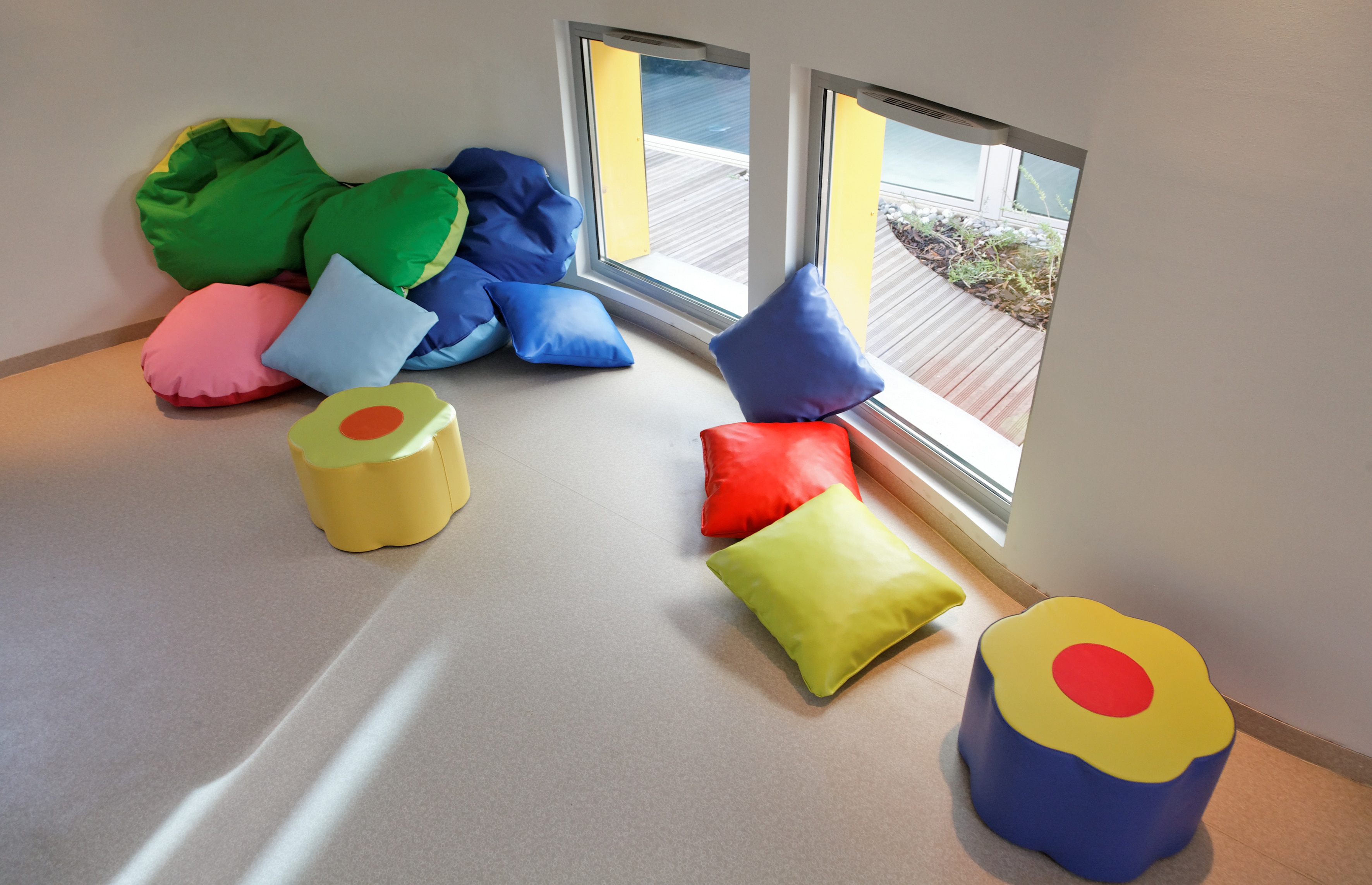 Salle de repos de la crèche : coussins colorés et poufs en forme de fleur au sol, grandes baies vitrées ouvertes sur la terrasse en bois